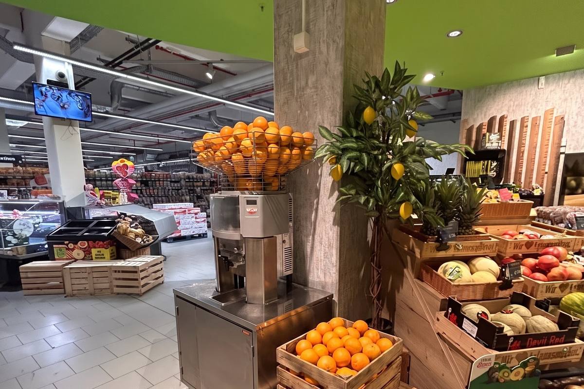 Die Obst- und Gemüseabteilung: die Saftpresse mit Orangen im Trichter, daneben ein Zitronenbäumchen und Holzkisten mit Melonen, Ananas und Äpfeln. (KI-modifizierte Darstellung)