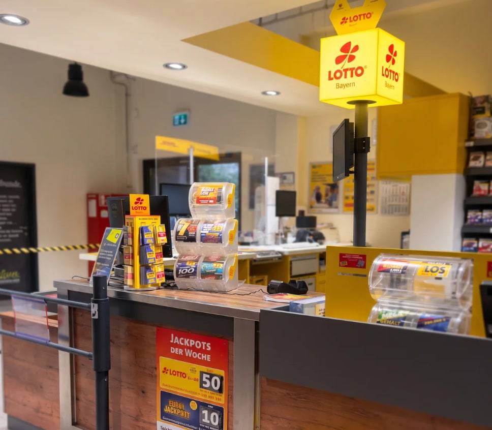 Die LOTTO-Annahmestelle im Markt: der beleuchtete LOTTO-Bayern-Würfel über dem Serviceplatz, davor die Spender mit Rubbellosen.