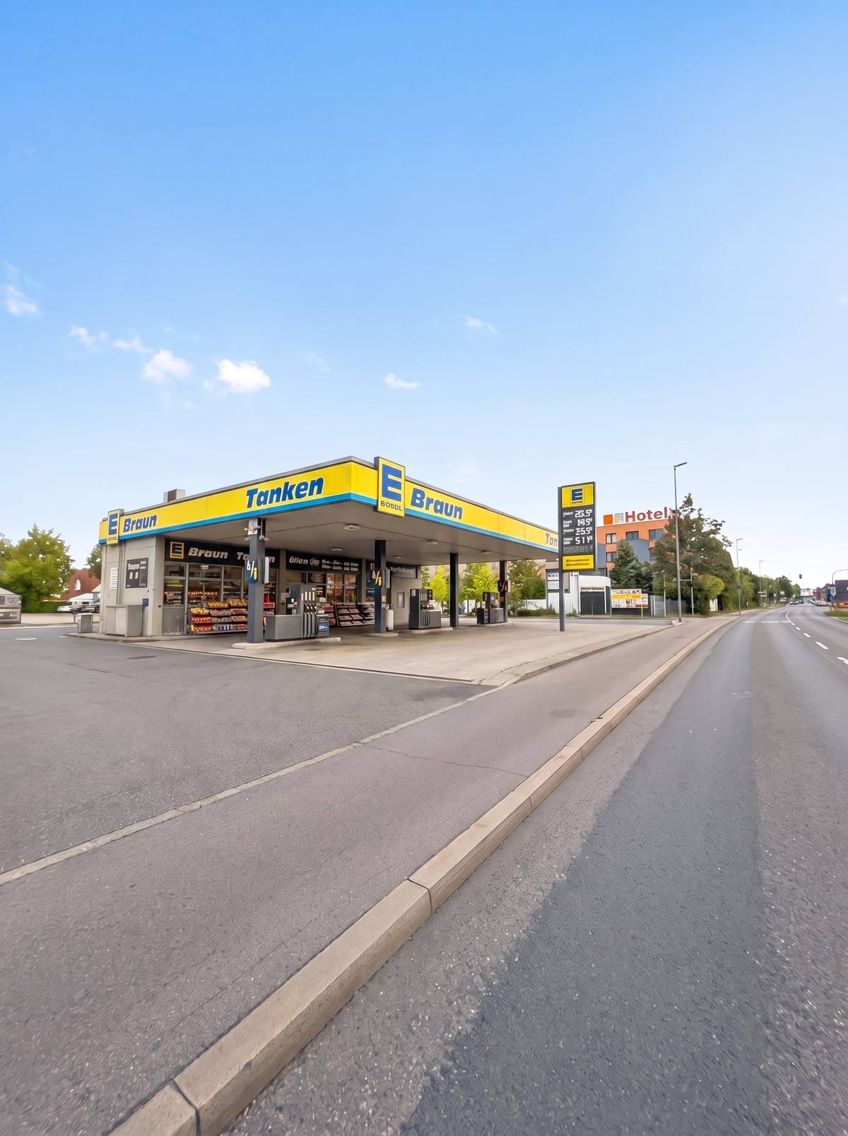 Die Tankstelle des Marktes bei Sonne: das gelb-blaue Vordach mit dem Schriftzug „Tanken“ über den Zapfsäulen, rechts die Preisstele, daneben die Straße. (KI-modifizierte Darstellung)