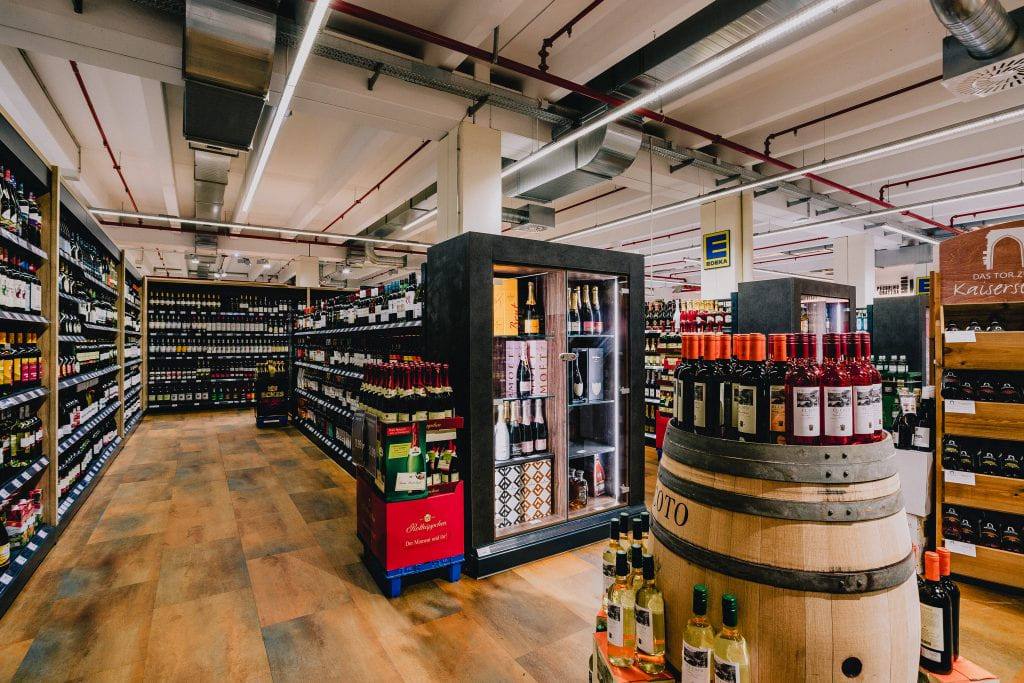Die Weinabteilung: Flaschen in Holzregalen rund um ein aufrecht stehendes Weinfass, das als Präsentationstisch dient.