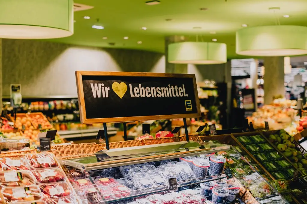 Das gerahmte Schild „Wir lieben Lebensmittel“ mit gelbem Herz über der Obstauslage, dahinter die grüne Decke der Markthalle.